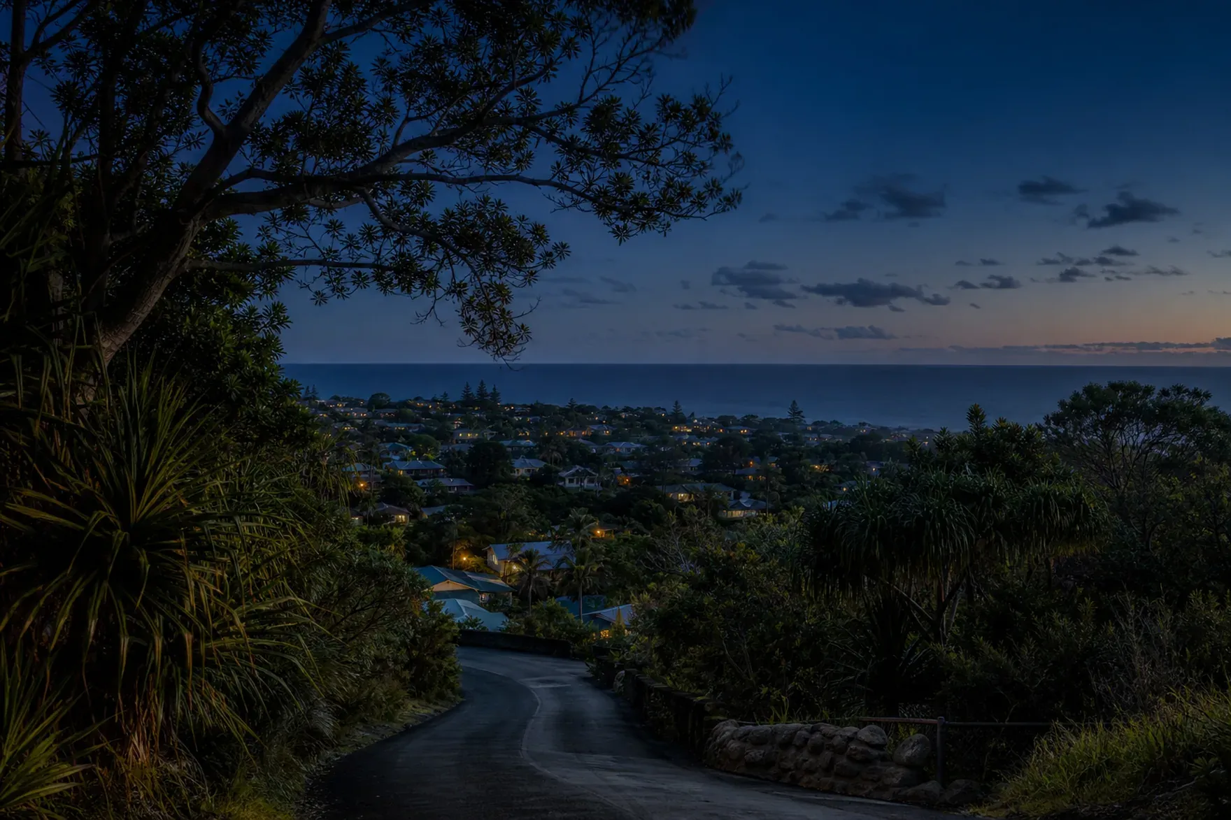 A hillside road above a coastal town at dusk, scattered lit windows among the trees. (AI-generated image)