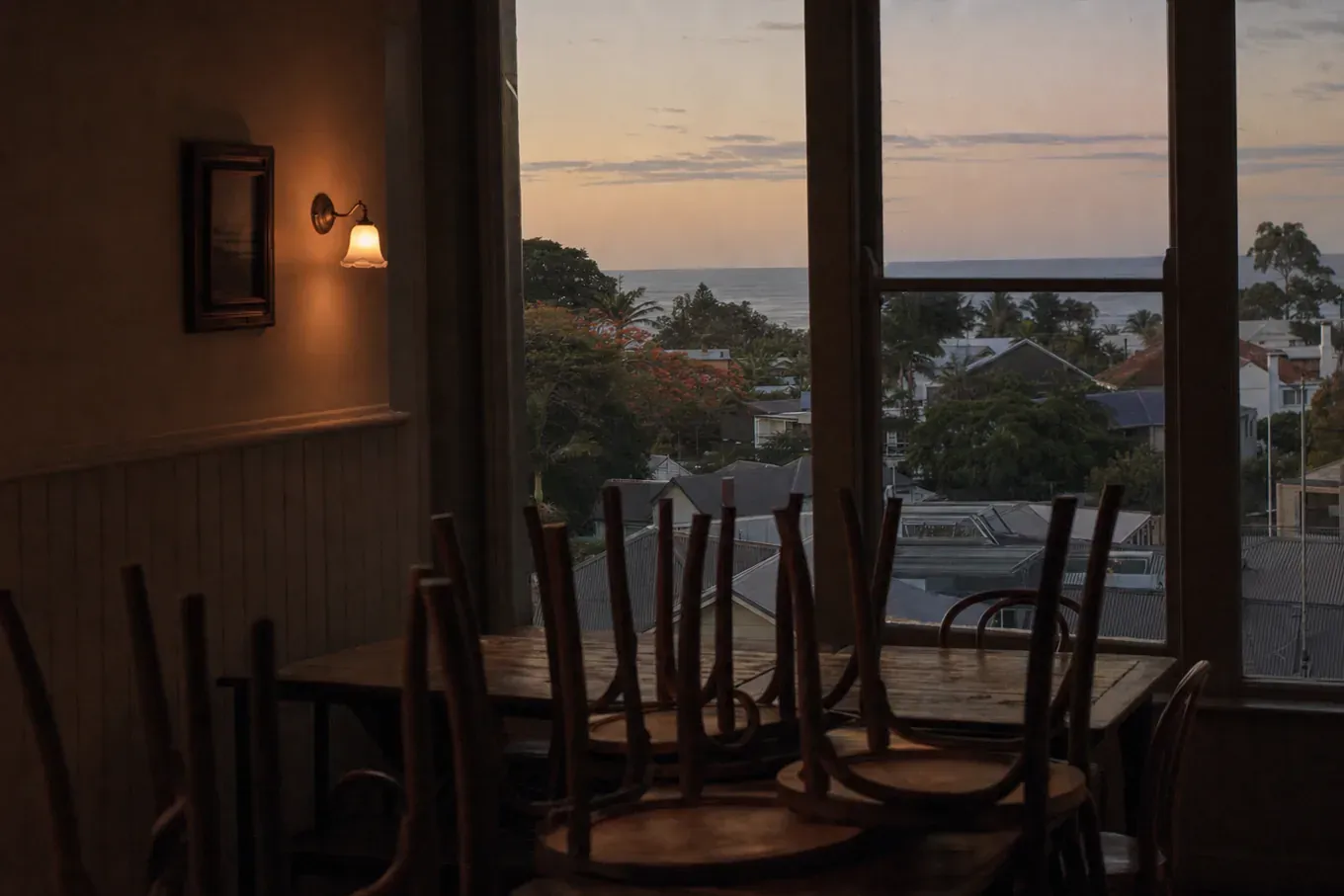 An empty upstairs dining room at dusk, chairs turned up on the tables, looking out over low tin roofs and palms to the sea. (AI-generated image)
