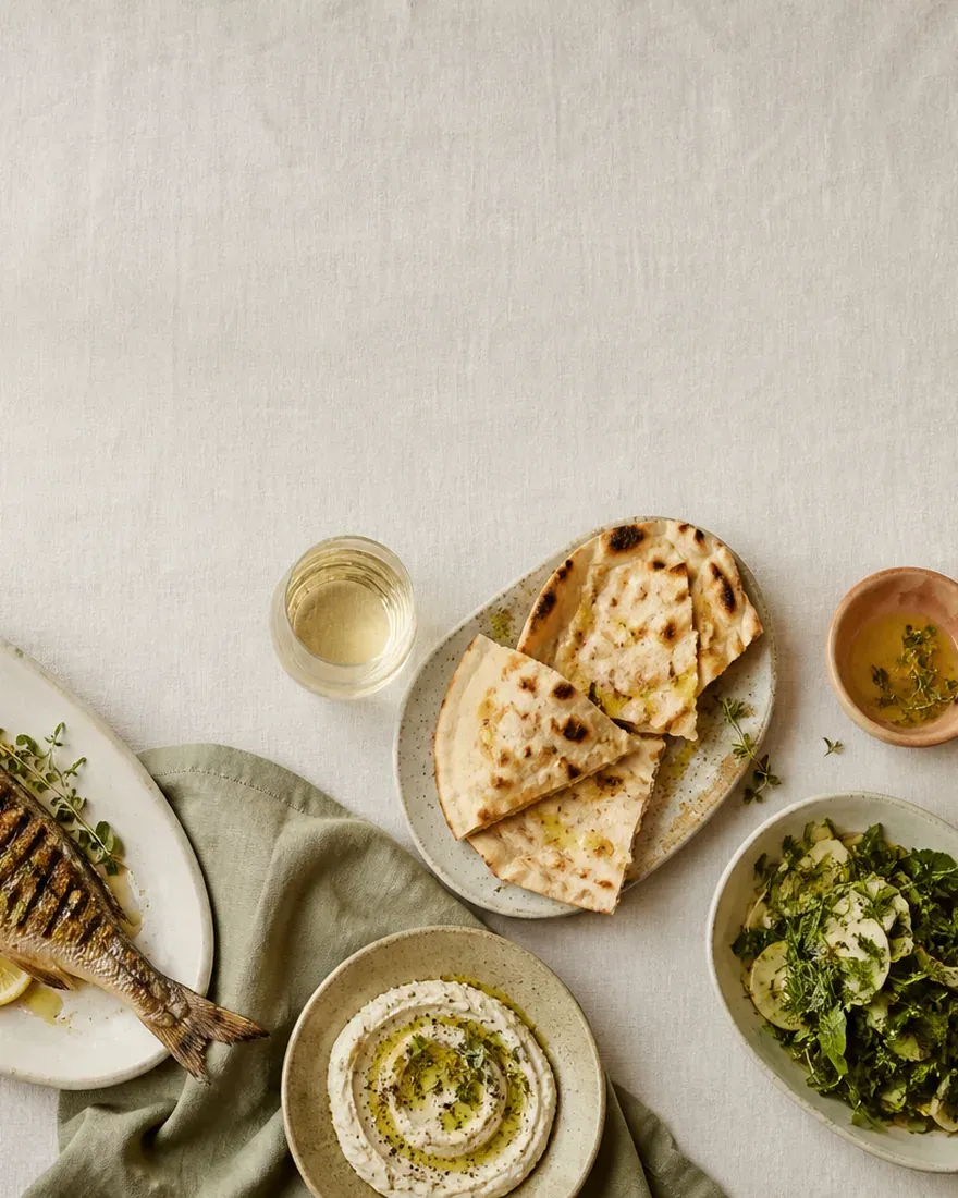 Shared Mediterranean plates — grilled fish, charred greens, flatbread and olives — on a linen table, seen from above. (AI-generated image)